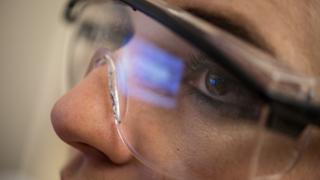 A close up on a researcher's face as she's watching a computer monitor during an experiment. The refection of the monitor can be seen in her safety goggles.